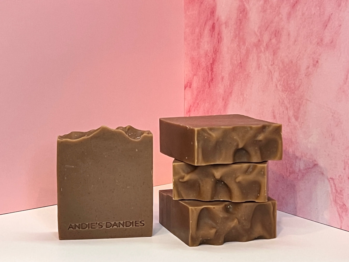 Three bars of brown soap stacked and one standing face forward on a white surface with 'Andie's Dandies' branding on a pink marble background