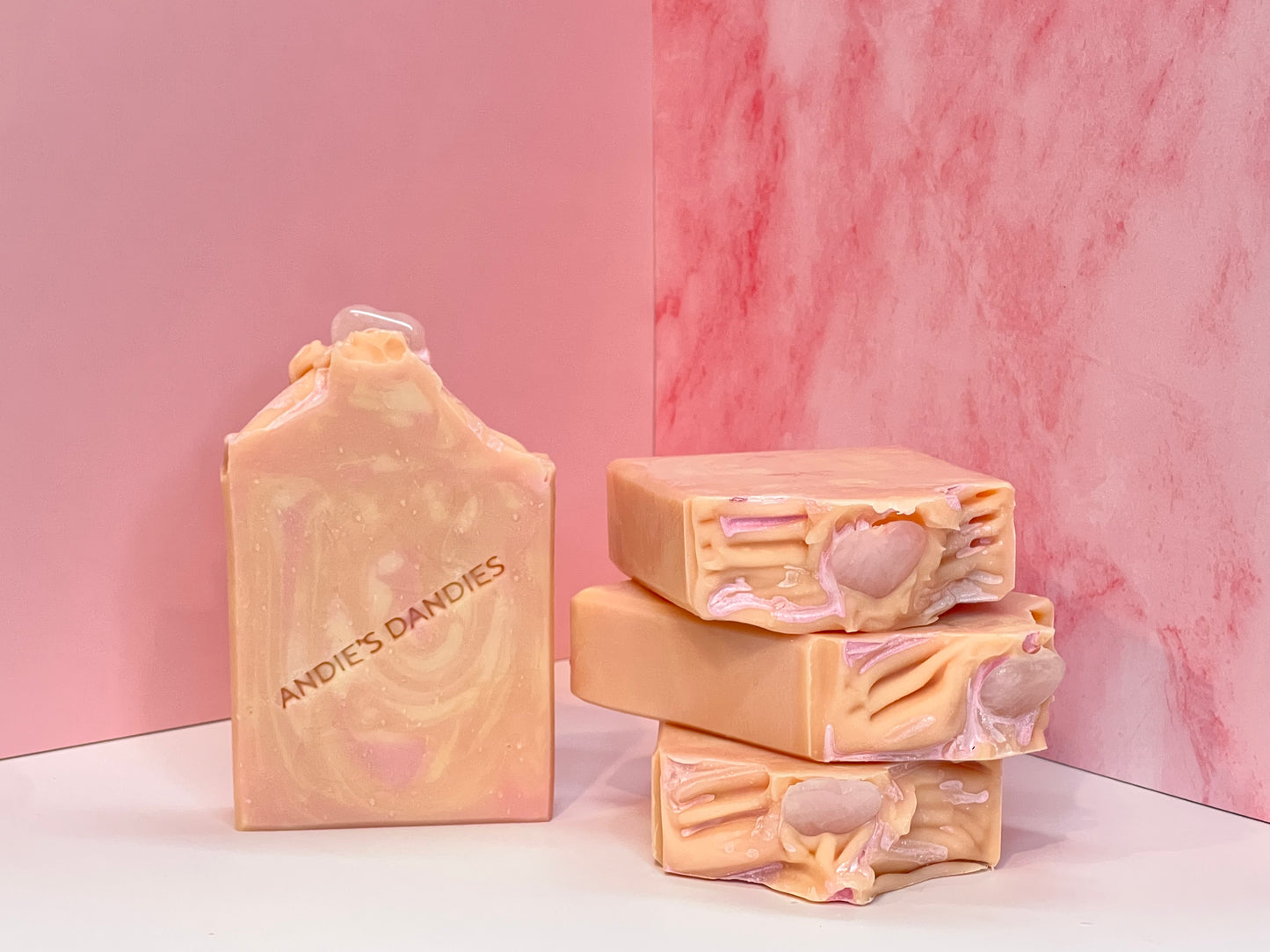 Stack of 3 pink and beige marbled soap bars with one forward facing with heart shaped rose quartz stones on top, with "Andie's Dandies" branding on a white surface with a pink marble background