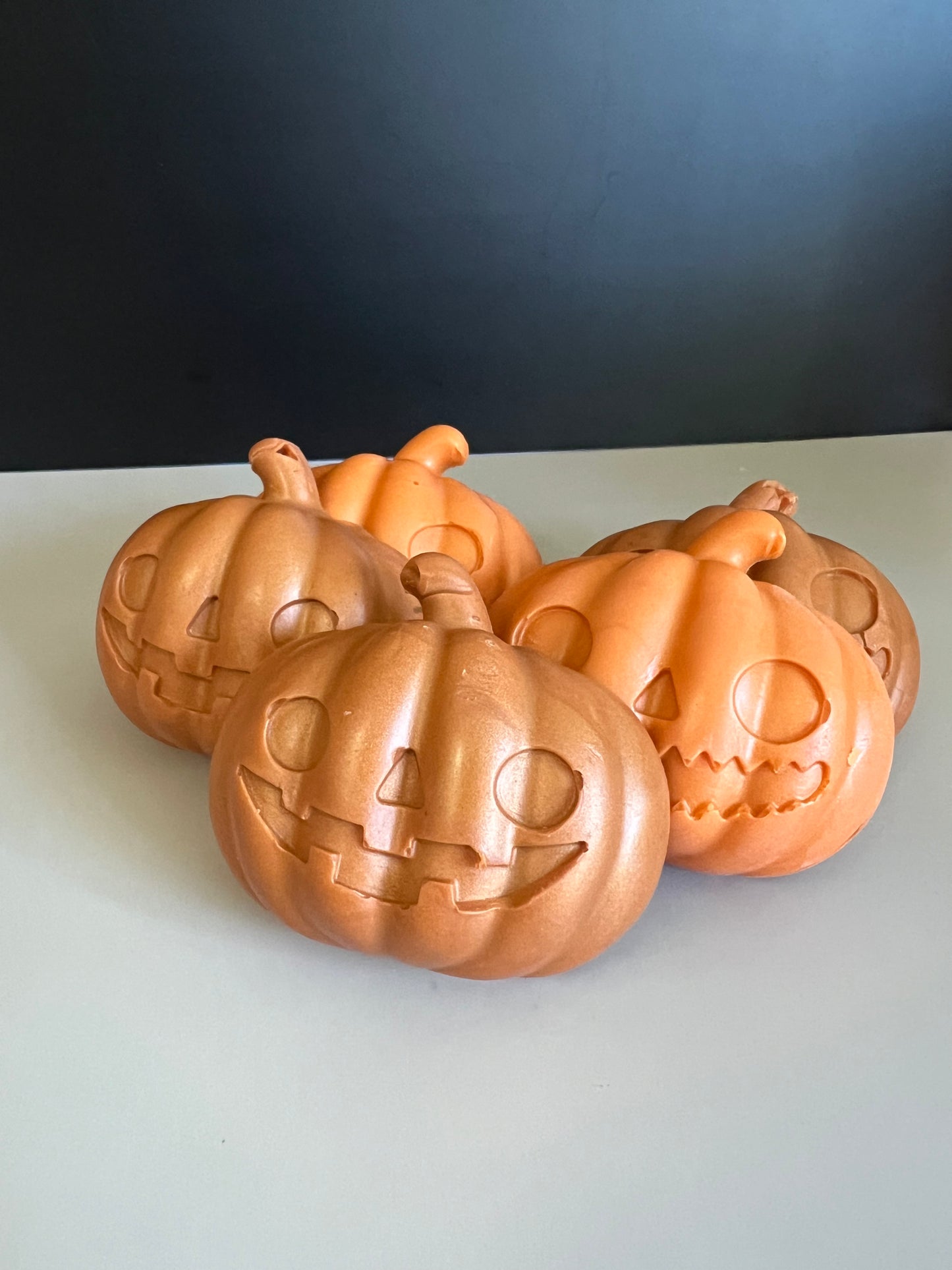 Five orange and orange-gold jack-o'-lantern pumpkin soaps with carved faces, arranged together on a light gray surface against a dark gray background.