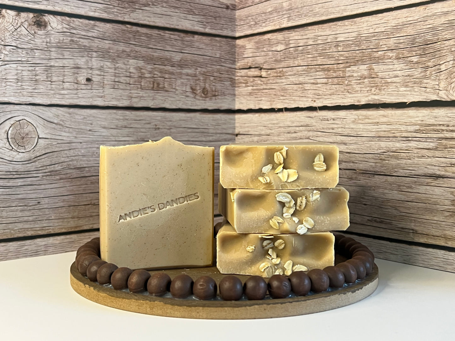 Four natural handmade soap bars displayed on a wooden stand against a rustic wood plank background. The face of the bar standing up is embossed with the brand name 'ANDIE'S DANDIES', and three others are stacked, decorated with visible oats on top. The soap is light tan with darker brown speckles from the colloidal oats.