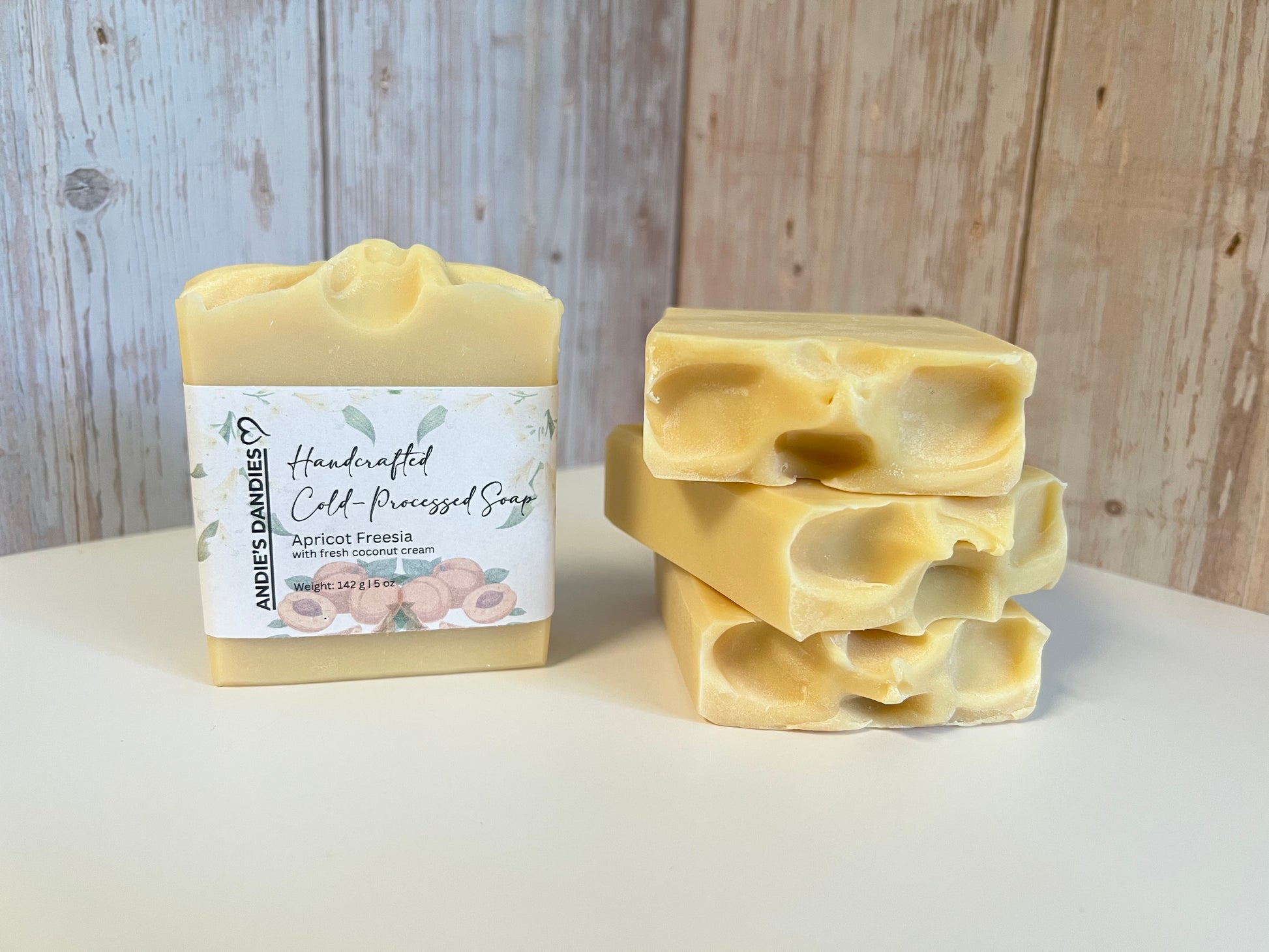 Handmade cold-processed soap bars in a pale apricot-yellow color, with one wrapped bar showing a label that reads 'Andie's Dandies' and three unwrapped bars stacked on the right, set against a rustic light wood background.