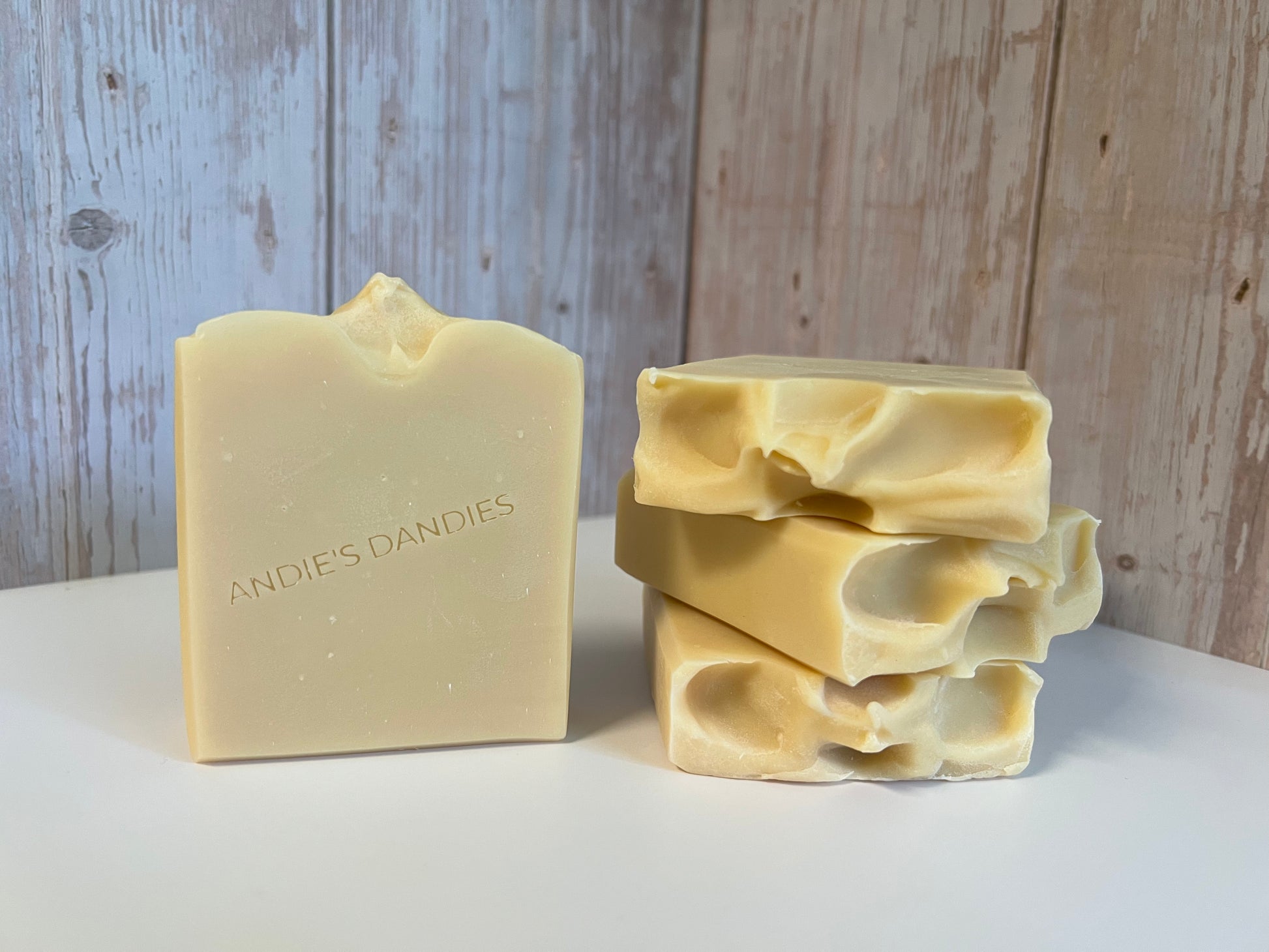 A handmade bar of soap in a warm cream color with the brand name 'ANDIE'S DANDIES' embossed on the front, standing next to a stack of three matching rough-cut soap bars on a white surface against a rustic wood-paneled background.