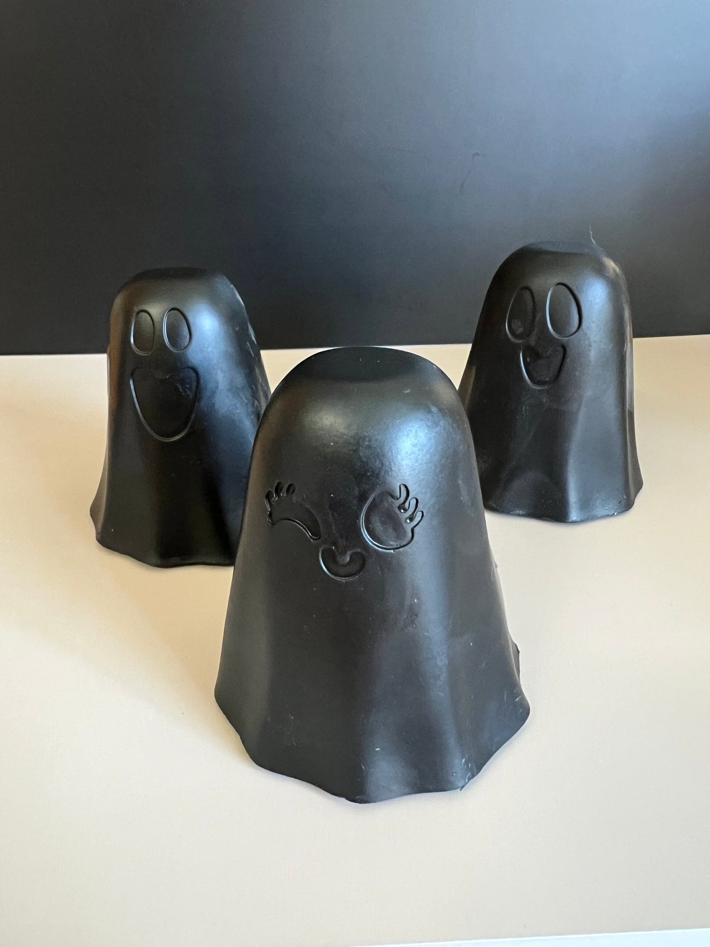 Three black ghost-shaped decorative soaps with carved facial features, arranged on a light surface against a dark gray background.