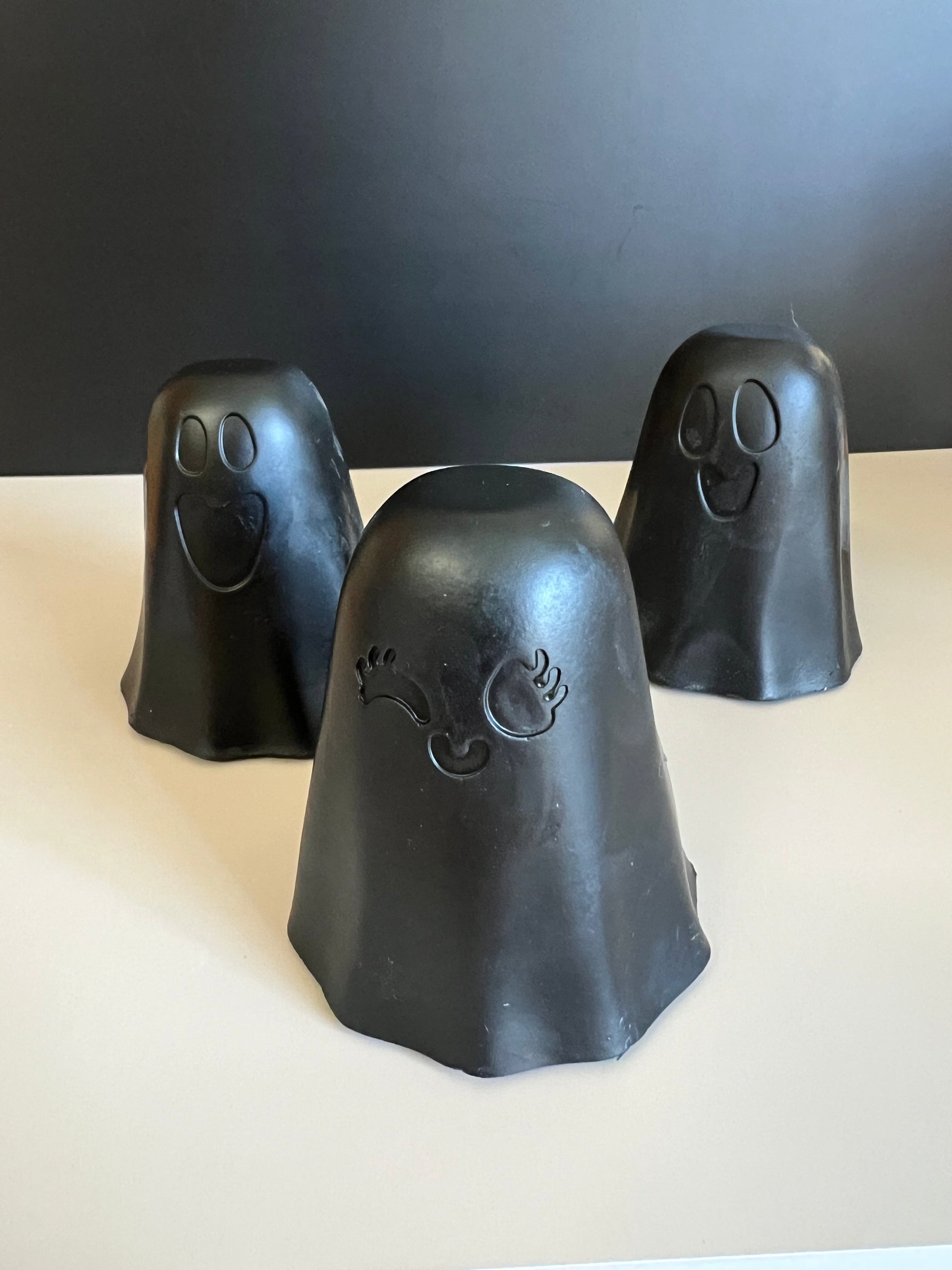 Three black ghost-shaped decorative soaps with carved facial features, arranged on a light surface against a dark gray background.