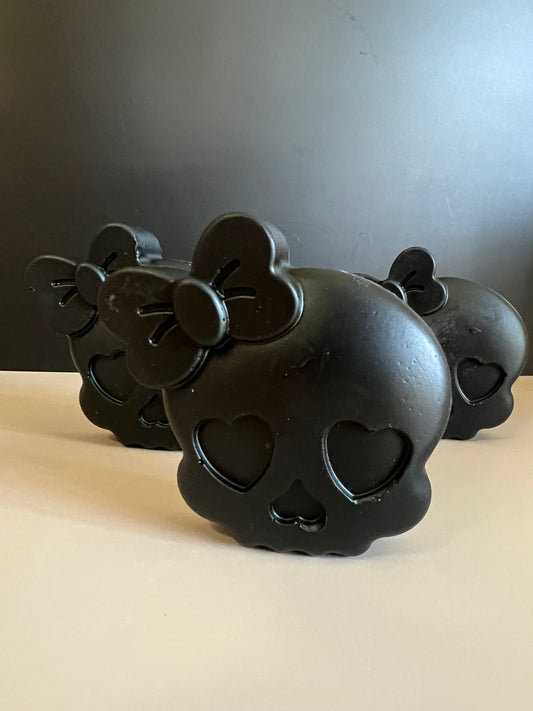 Three black skull-shaped soaps with heart-shaped eye cutouts and bow details, arranged on a light beige surface against a dark gray background.