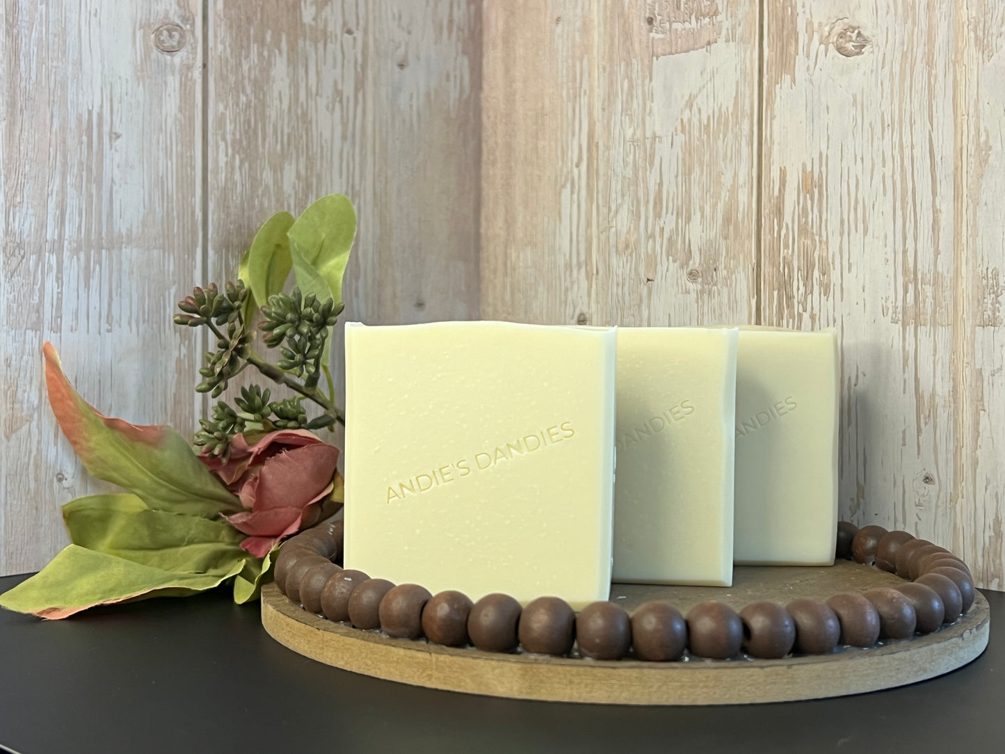 Three rectangular bars of pale cream soap standing upright on a round wooden tray, styled with dried flowers and green leaves. The soap is embossed with the brand name 'ANDIE'S DANDIES'.