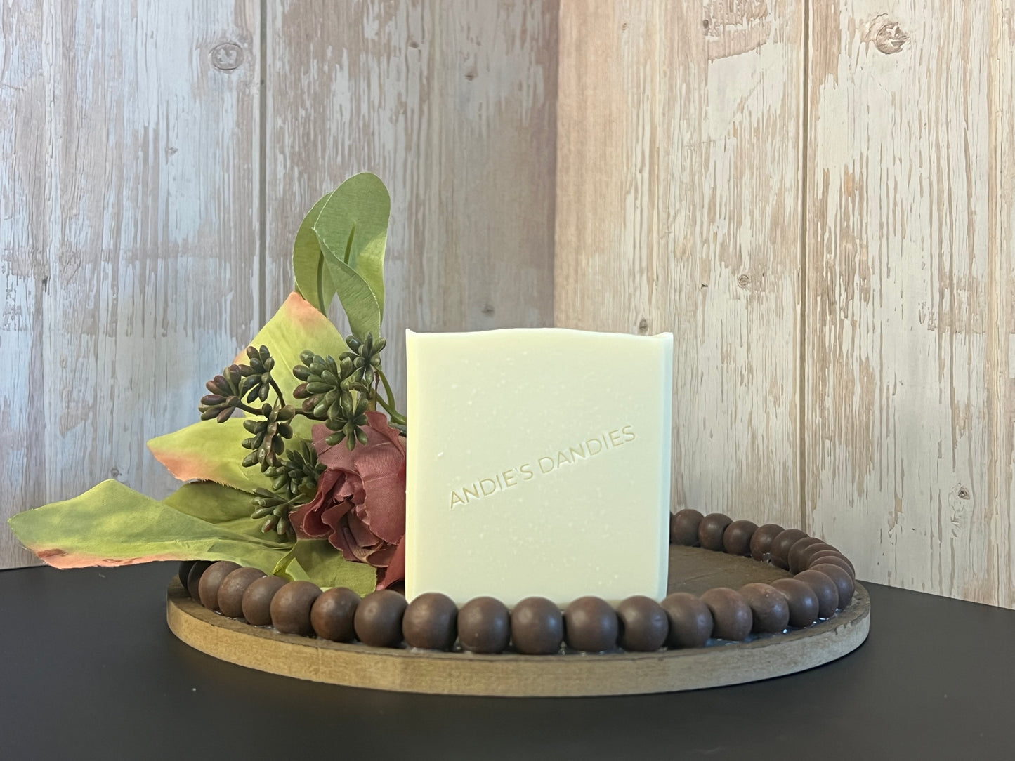 A rectangular bar of pale cream soap standing upright on a round wooden tray, styled with dried flowers and green leaves. The soap is embossed with the brand name 'ANDIE'S DANDIES'.