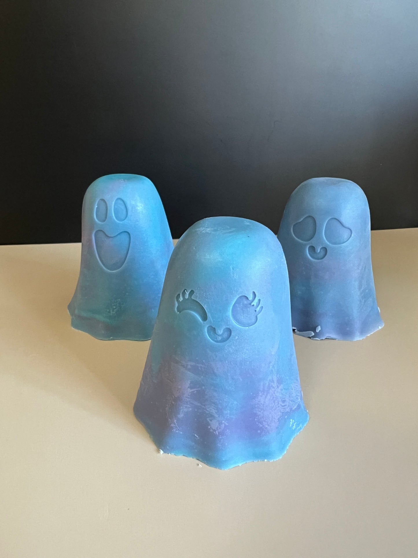 Three ghost-shaped soaps in shades of blue and purple, featuring cute carved out facial features, arranged on a light beige surface against a dark gray background.