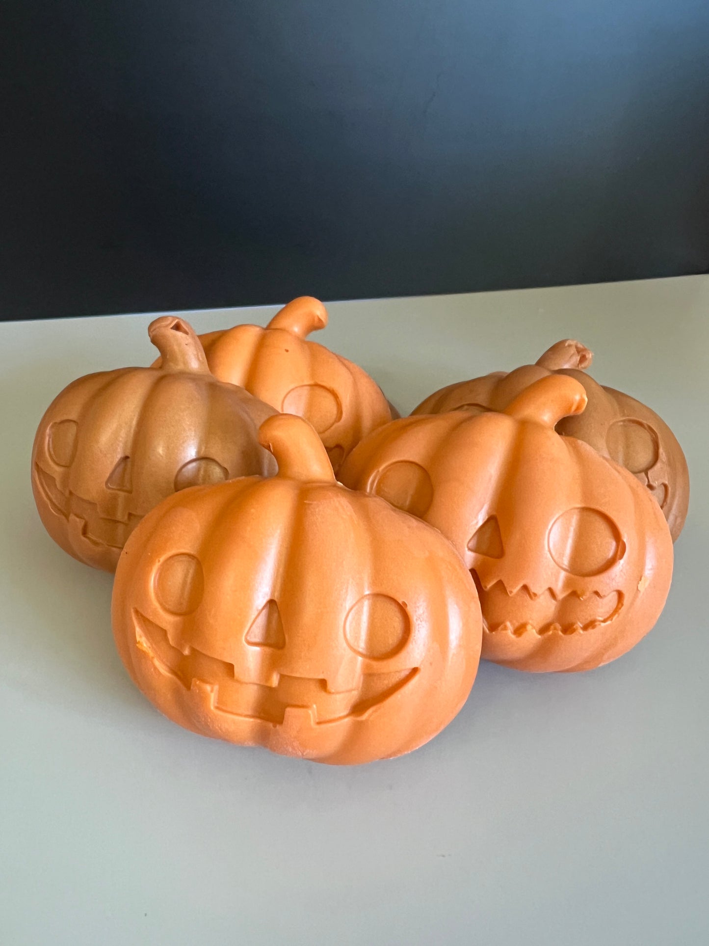 Five orange jack-o'-lantern pumpkin soaps with carved faces, arranged together on a light gray surface against a dark gray background.