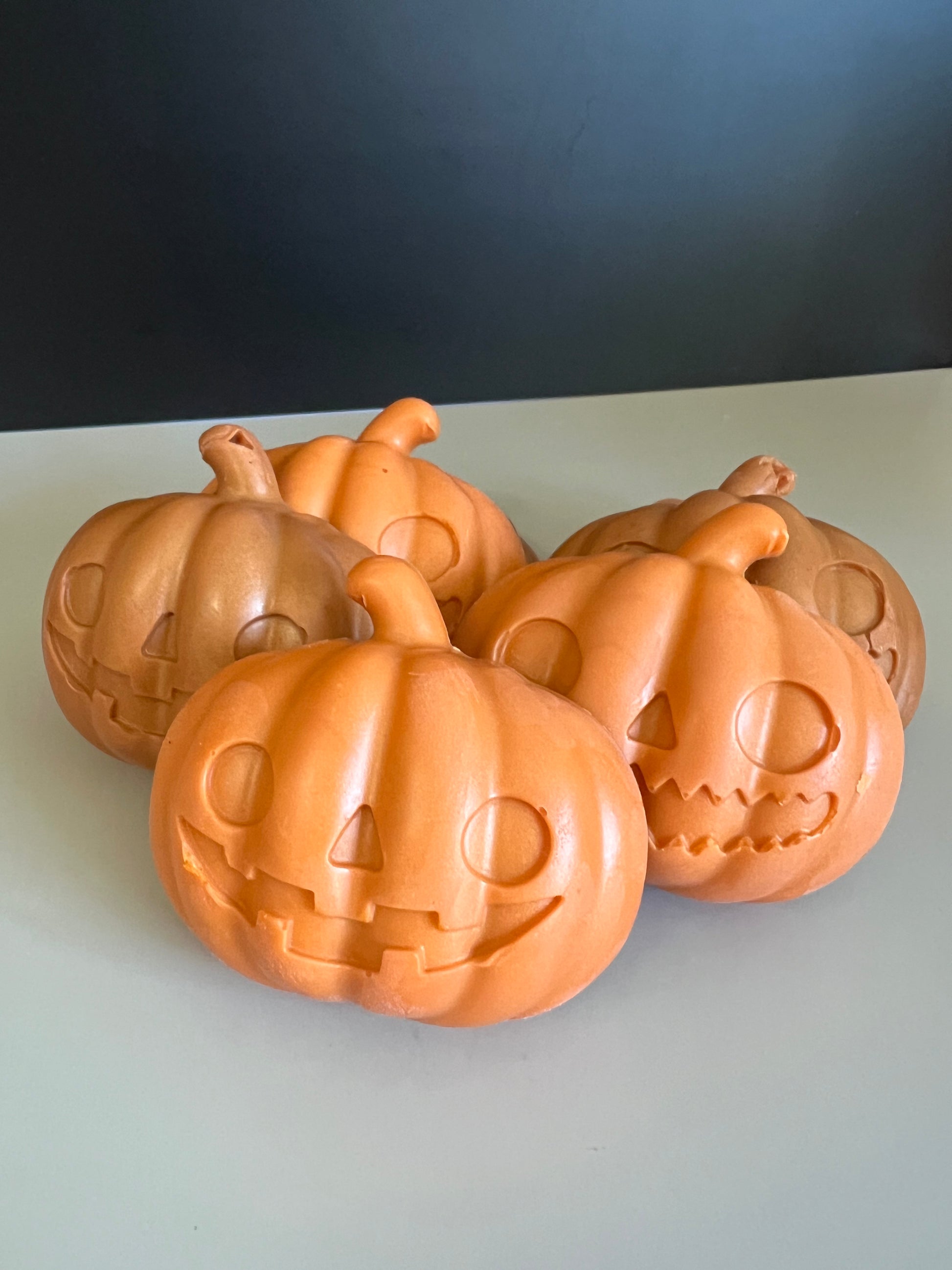 Five orange jack-o'-lantern pumpkin soaps with carved faces, arranged together on a light gray surface against a dark gray background.