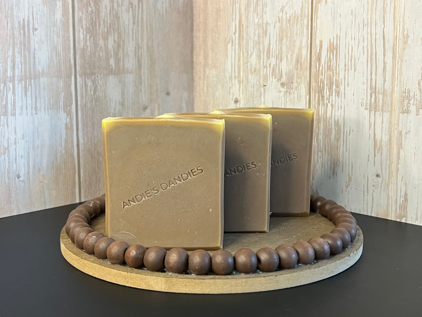 Three brown rectangular bars of soap labeled 'ANDIE'S DANDIES' standing upright on a brown round wooden bead tray, set against a light wood plank wall.