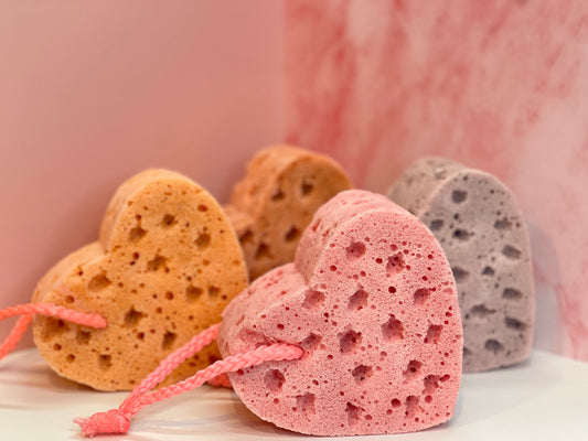 Heart-shaped, soap-infused sponges in various colors on a white surface against a pink marbled background. Sponge colors shown are pink, orange, muted purple, and a darker orangey-pink. 