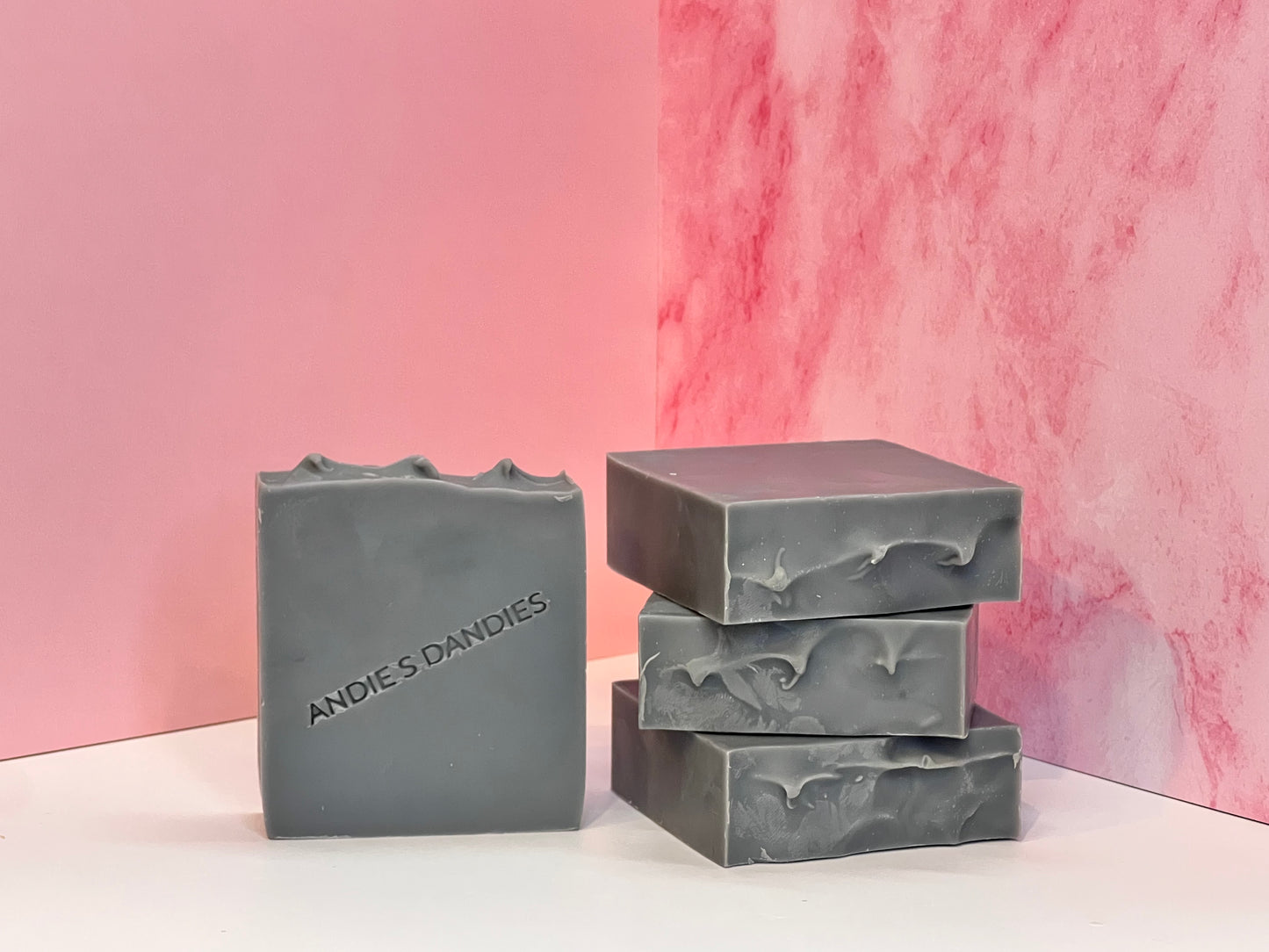 Three bars of grey soap stacked and one standing face forward on a white surface with 'Andie's Dandies' branding on a pink marble background