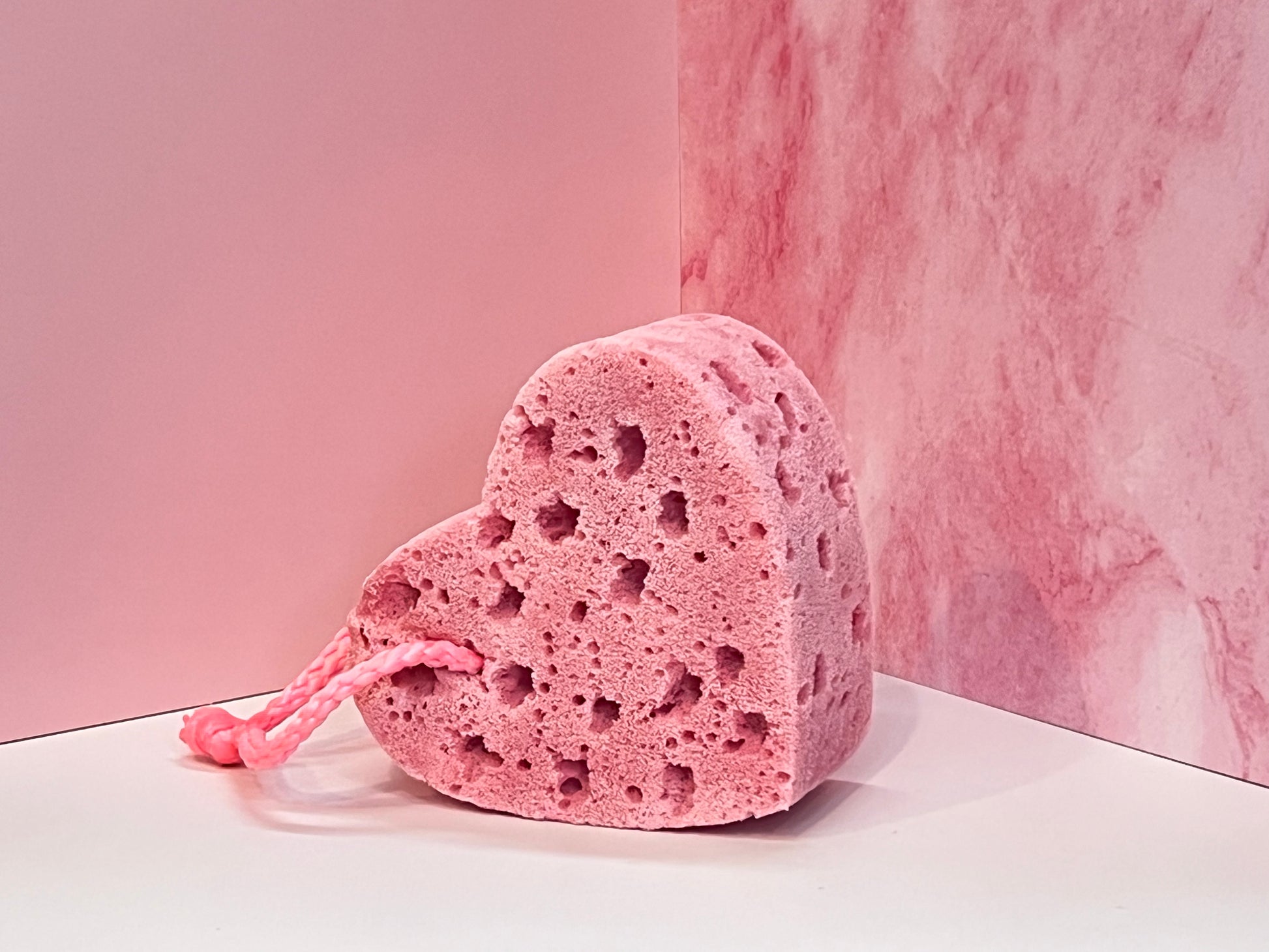 Heart-shaped, pink soap-infused sponge with pink string on a white surface against a pink marble background