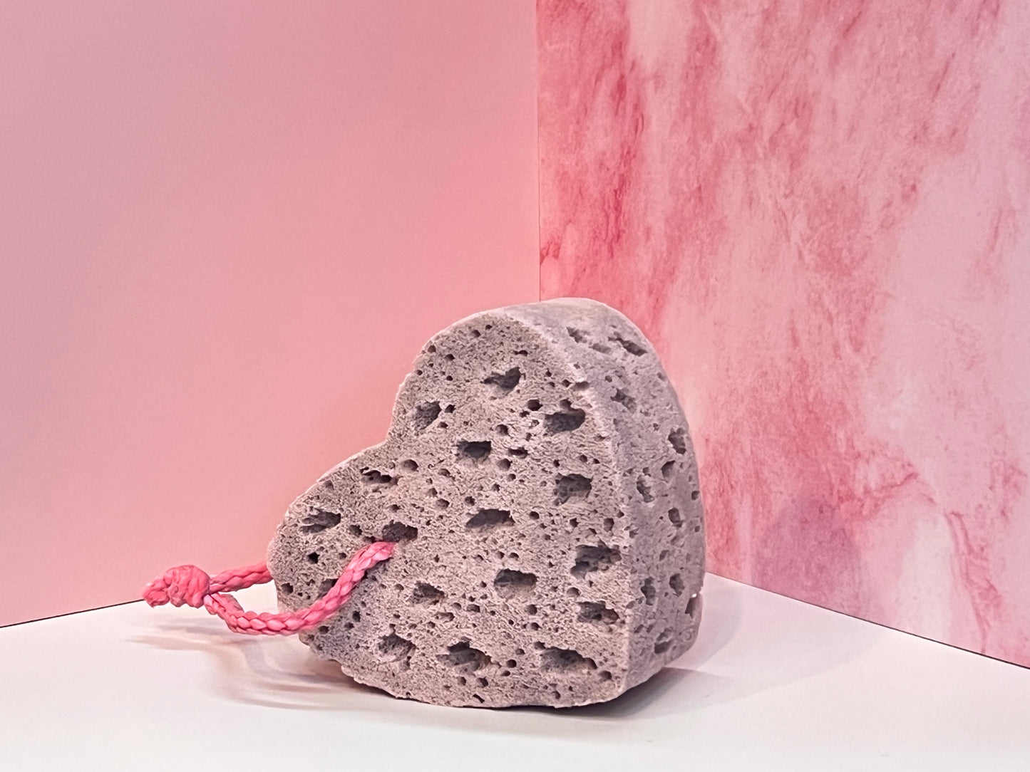 Heart-shaped, muted purple, soap-infused sponge with pink string on a white surface against a pink marble background