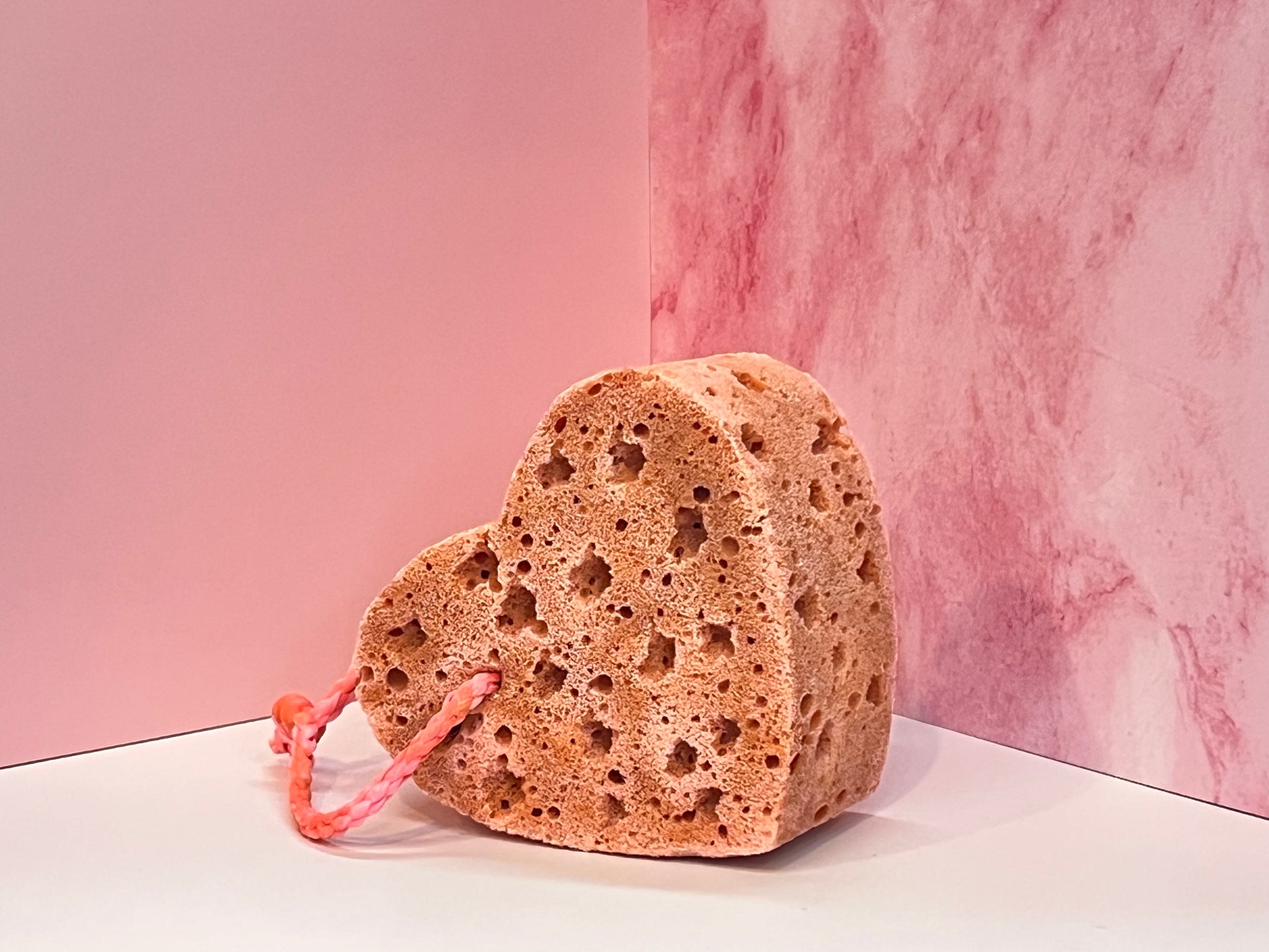 Heart-shaped, orangey-pink, soap-infused sponge with pink string on a white surface against a pink marble background
