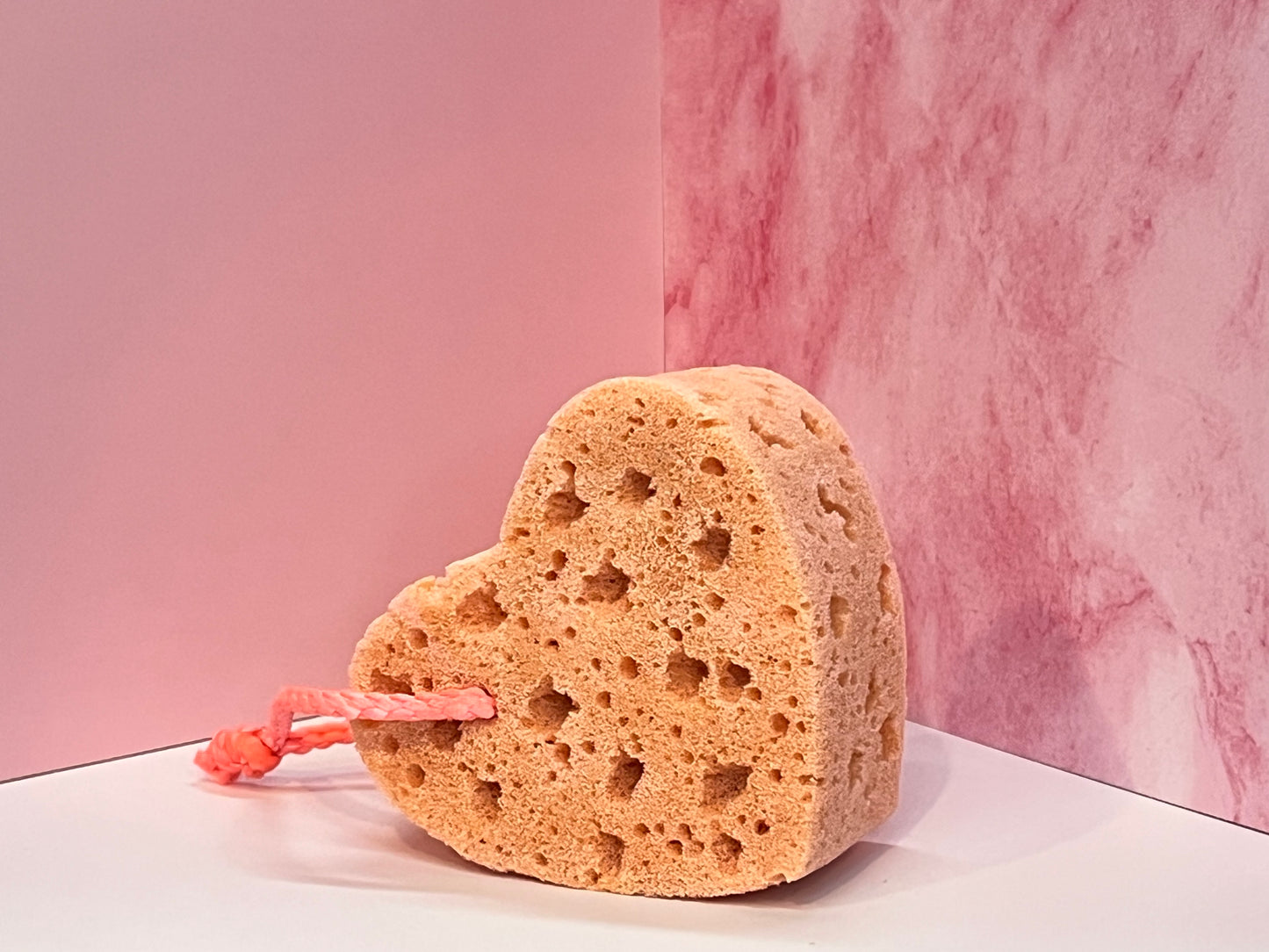 Heart-shaped, orange soap-infused sponge with pink string on a white surface against a pink marble background