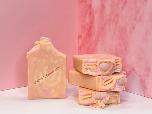 Stack of 3 pink and beige marbled soap bars with one forward facing with heart shaped rose quartz stones on top, with "Andie's Dandies" branding on a white surface with a pink marble background