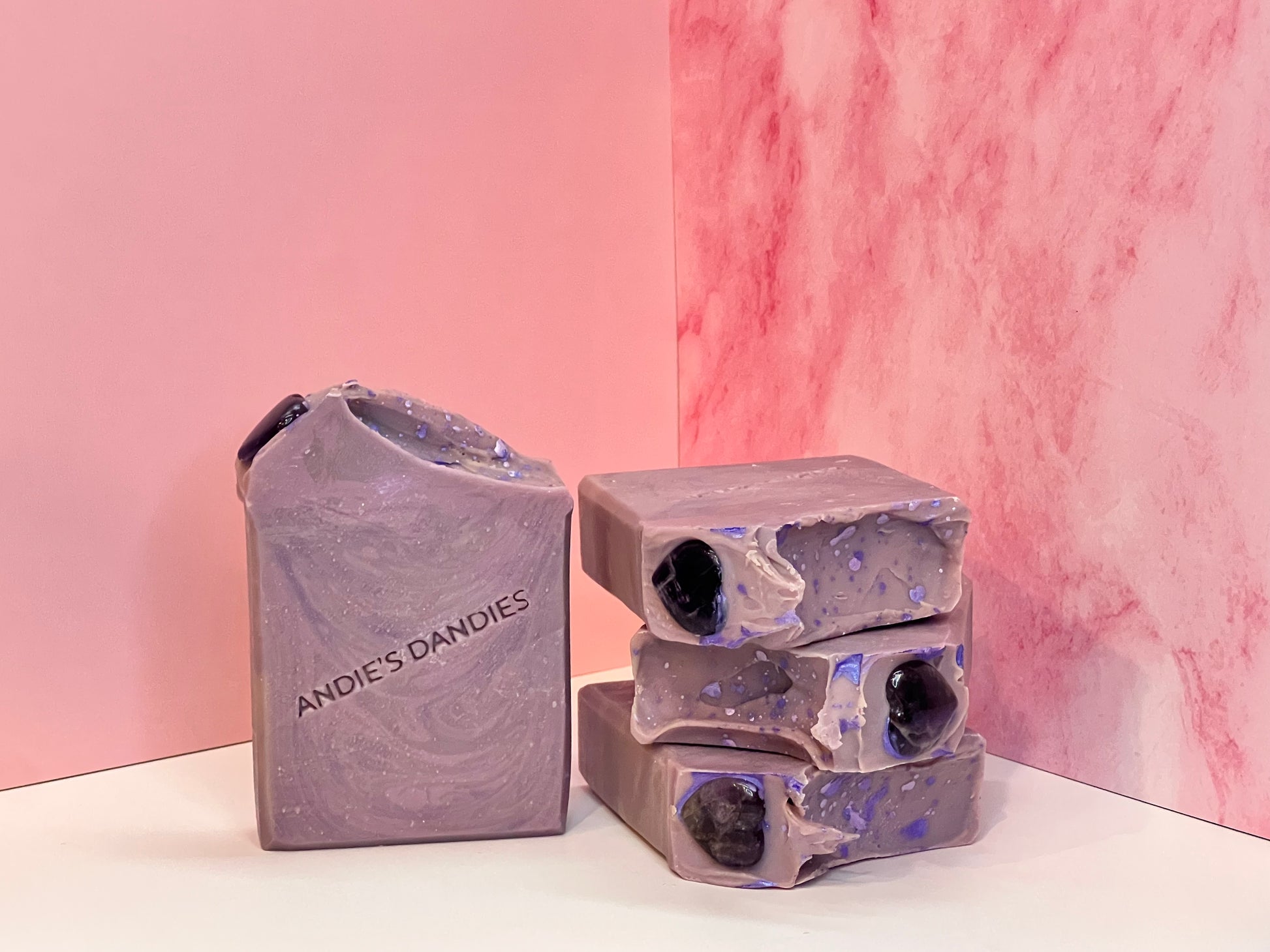 Stack of 3 purple marbled soap bars with one forward facing with heart shaped amethyst stones on top, with "Andie's Dandies" branding on a white surface with a pink marble background