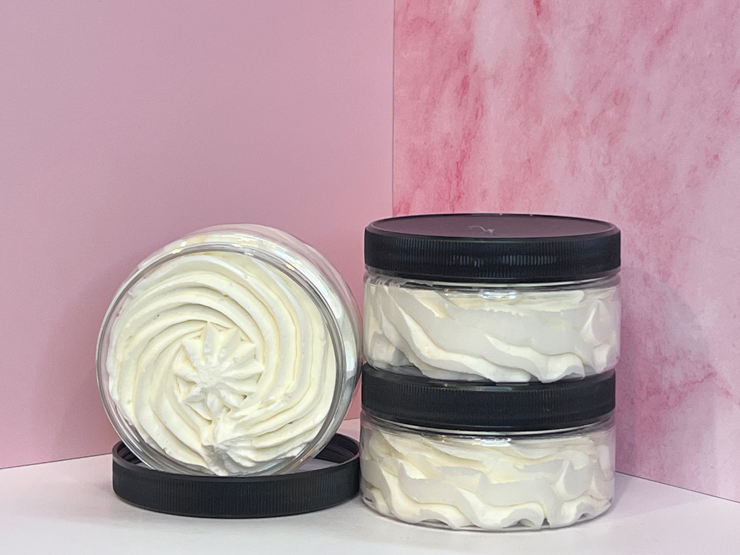 Two shallow wide mouth straight sided jars with black ribbed tops stacked, and one open sitting on its side in its lid, showing the whipped body botter contents that resemble whipped cream, on a white surface with a pink marble background