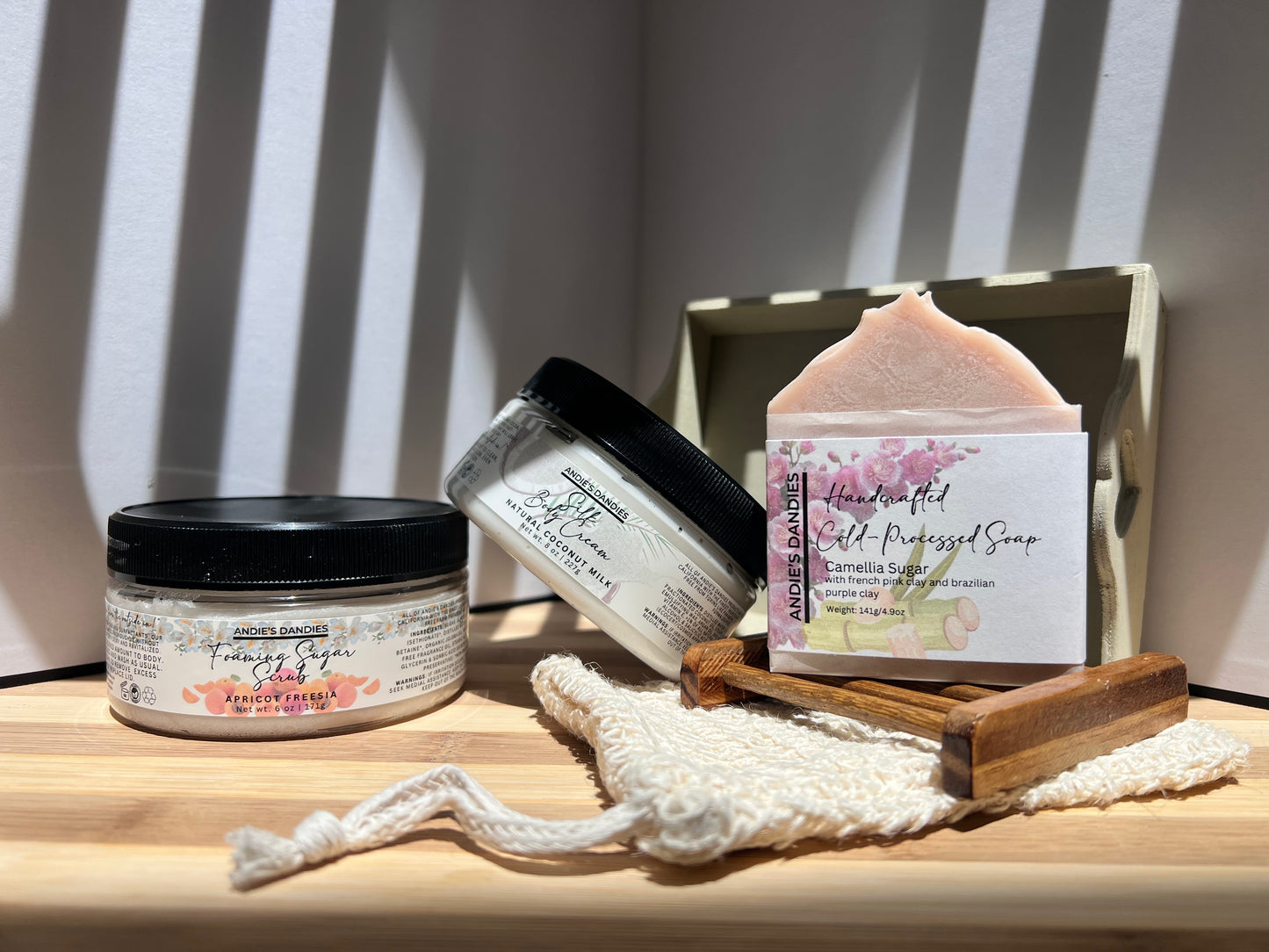 Assorted skincare products including jars of foaming sugar scrub, silk body crean and and a soap bar on a wooden soap tray and a sisal soap bag, on a wooden surface.