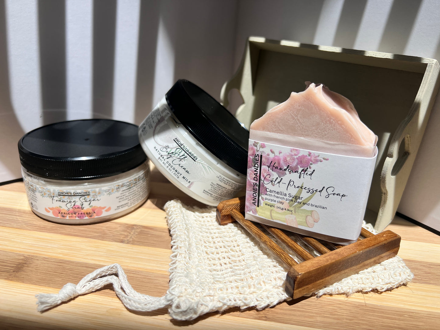 Assorted skincare products including jars of foaming sugar scrub, silk body crean and and a soap bar on a wooden soap tray and a sisal soap bag, on a wooden surface.