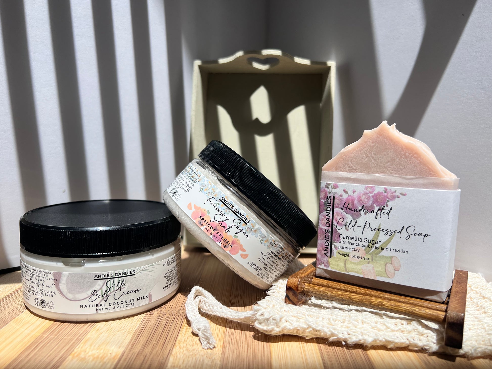 Assorted skincare products including jars of foaming sugar scrub, silk body crean and and a soap bar on a wooden soap tray and a sisal soap bag, on a wooden surface.