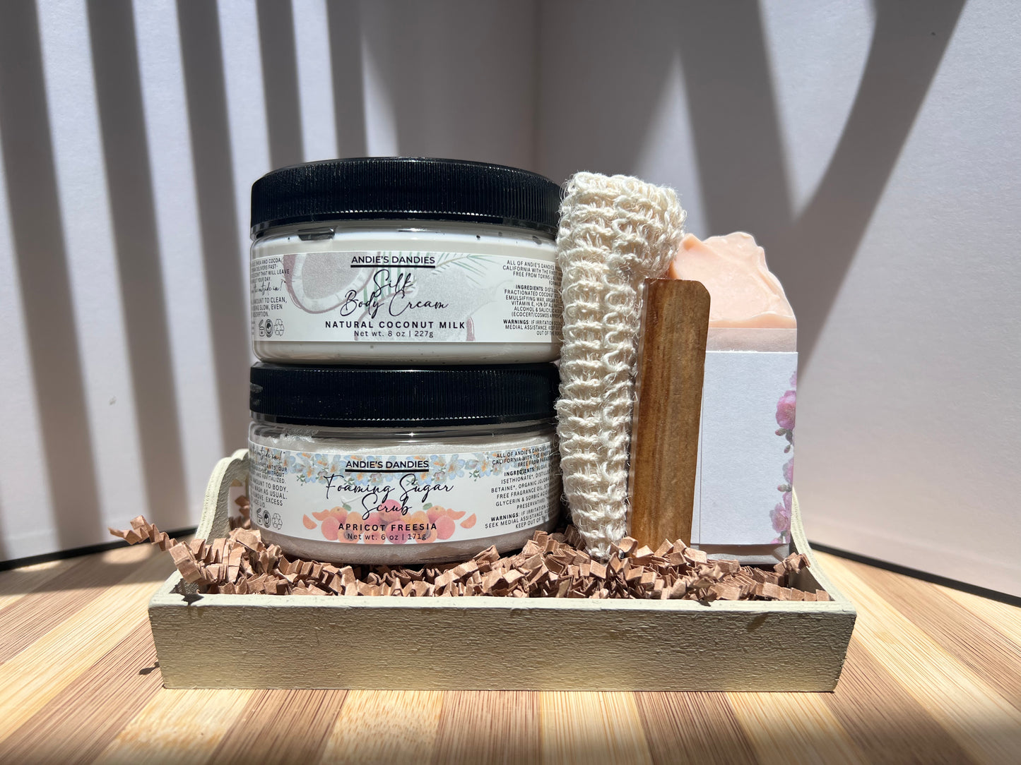 Assorted skincare products including jars of foaming sugar scrub, silk body crean and and a soap bar on a wooden soap tray and a sisal soap bag, on a wooden surface.