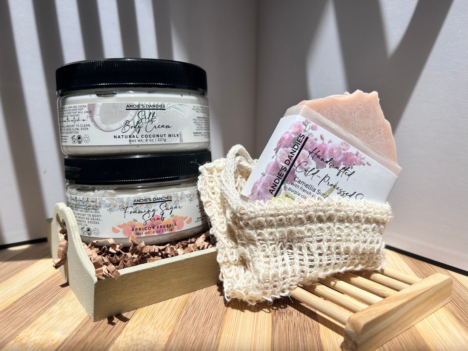 Assorted skincare products including jars of foaming sugar scrub, silk body crean and and a soap bar on a wooden soap tray and a sisal soap bag, on a wooden surface.
