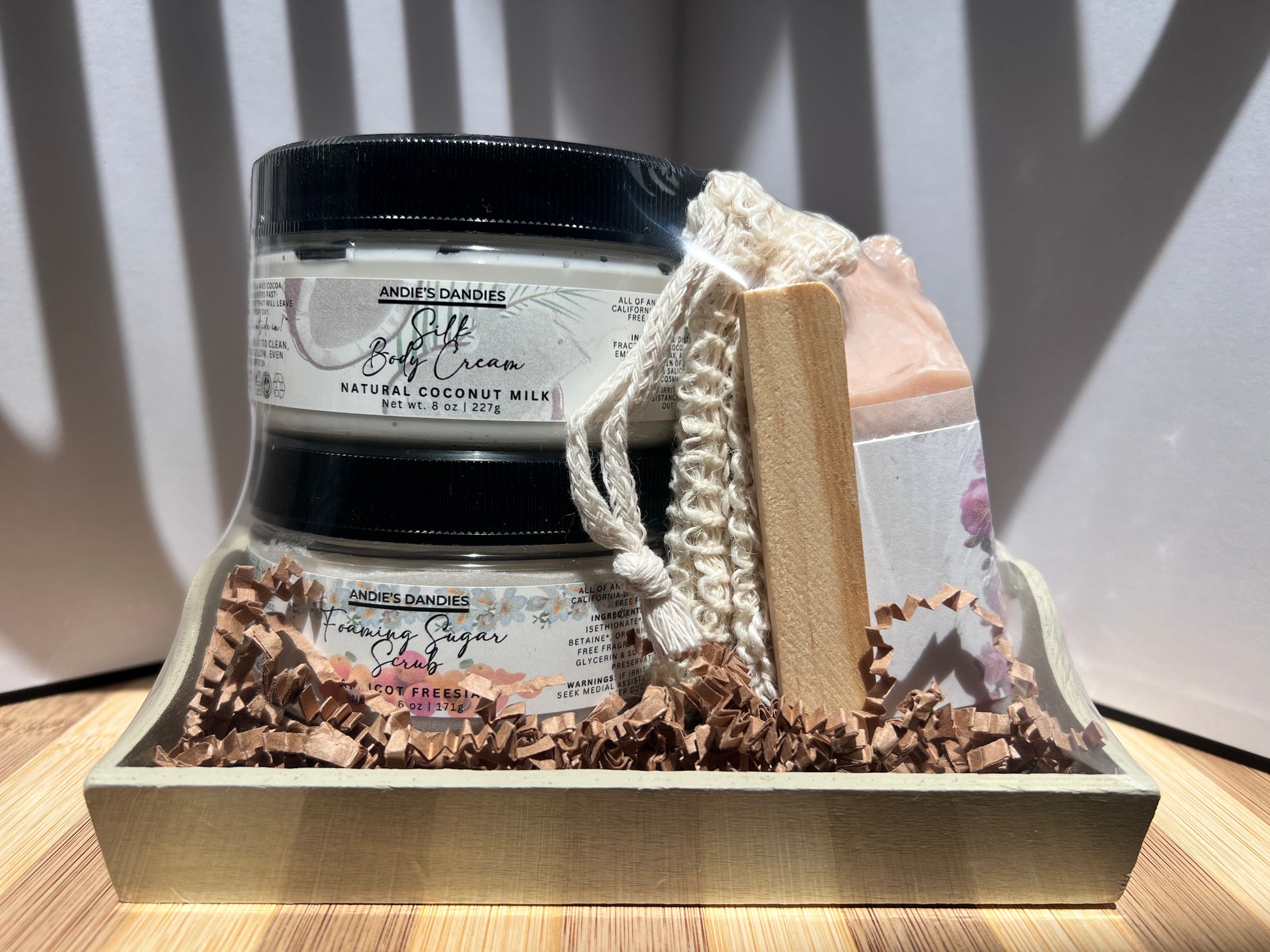 Assorted skincare products including jars of foaming sugar scrub, silk body crean and and a soap bar on a wooden soap tray and a sisal soap bag, shrink wrapped in a wooden tray.
