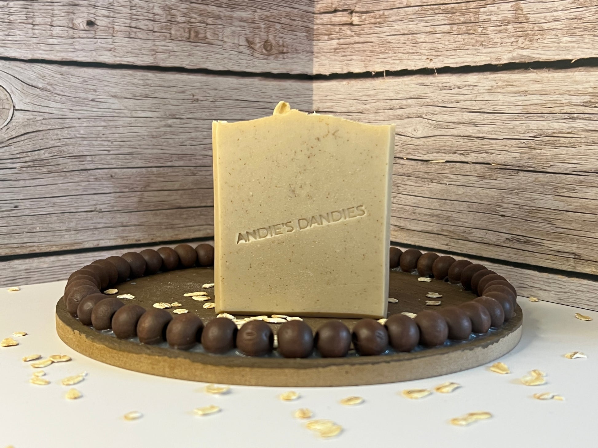 A rectangular bar of soap standing upright on a round wooden board, with dark brown beads around the edge. The soap is light tan with darker brown speckles from the colloidal oats, with the brand name 'ANDIE'S DANDIES' embossed on the front.