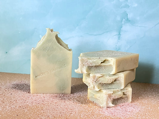 Three bars of very muted, pale yellow and green multicolored soap, stacked on a textured surface with a light blue background
