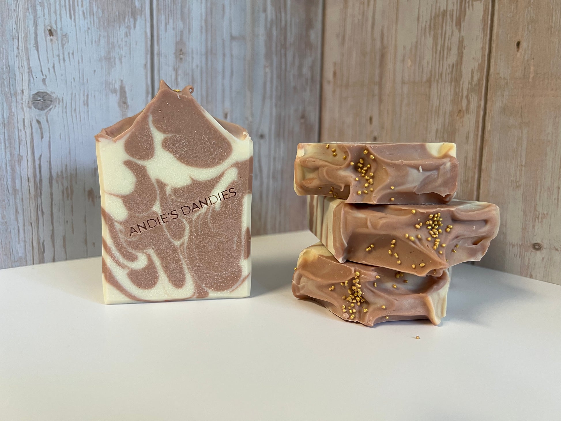 A handmade soap bar with a brown and cream swirl pattern and the brand name 'ANDIES DANDIES' printed on it, standing next to three stacked chocolate-dipped soap bars with gold sprinkles on a white surface against a rustic wood plank background.