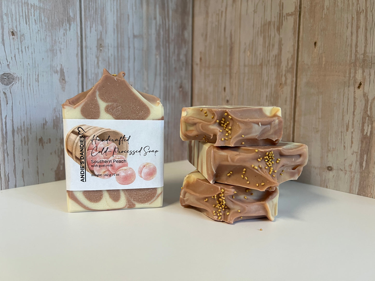 Handmade cold-pressed soap bars with a brown and cream swirl pattern, displayed on a white surface against a rustic light wood plank background. One soap bar stands upright with a white paper label with the brand name 'ANDIES DANDIES' printed on it, and three stacked soap bars are shown beside it, all featuring brown and cream swirls with small gold sprinkles.