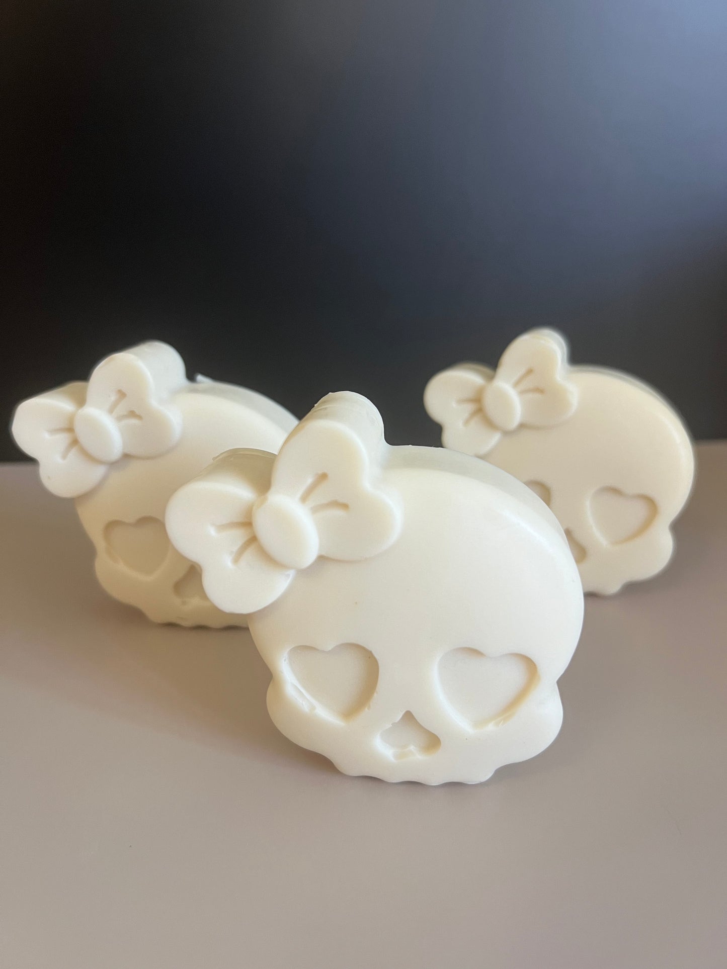 Three white skull-shaped decorative soaps with heart-shaped eye and nose cavities and a bow detail on top, arranged on a light surface against a dark background.