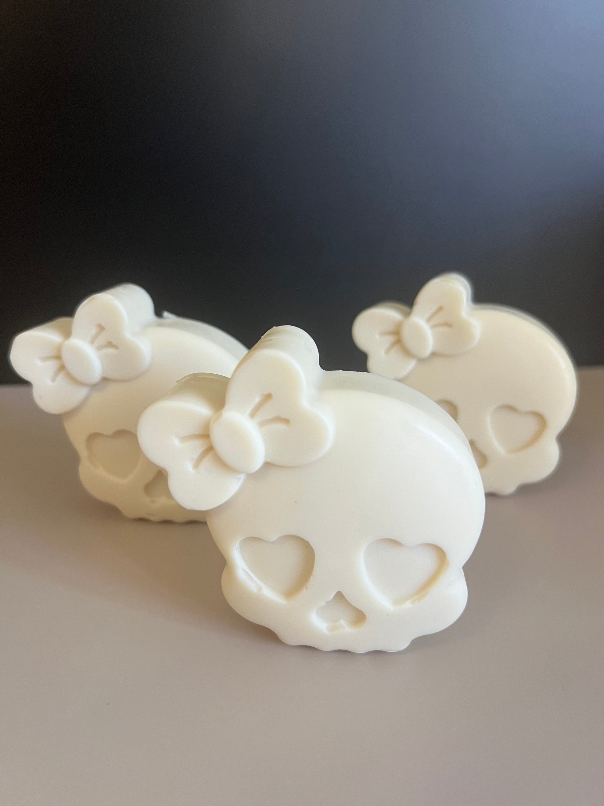 Three white skull-shaped decorative soaps with heart-shaped eye and nose cavities and a bow detail on top, arranged on a light surface against a dark background.