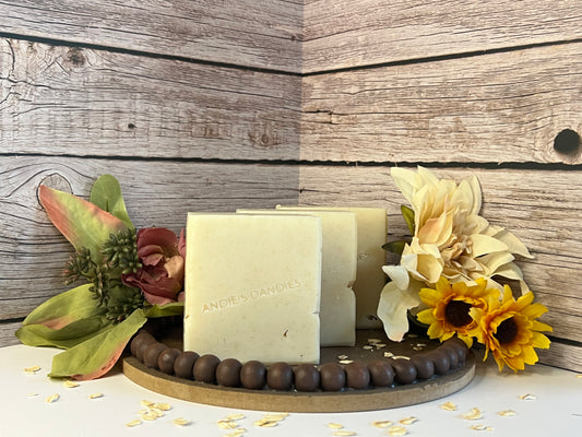 Three rectangular bars of soap standing upright on a round wooden tray, styled with flowers and oats against a rustic wood plank background. The soap has a light cream color and the brand name 'ANDIE'S DANDIES' is embossed on the front bar.