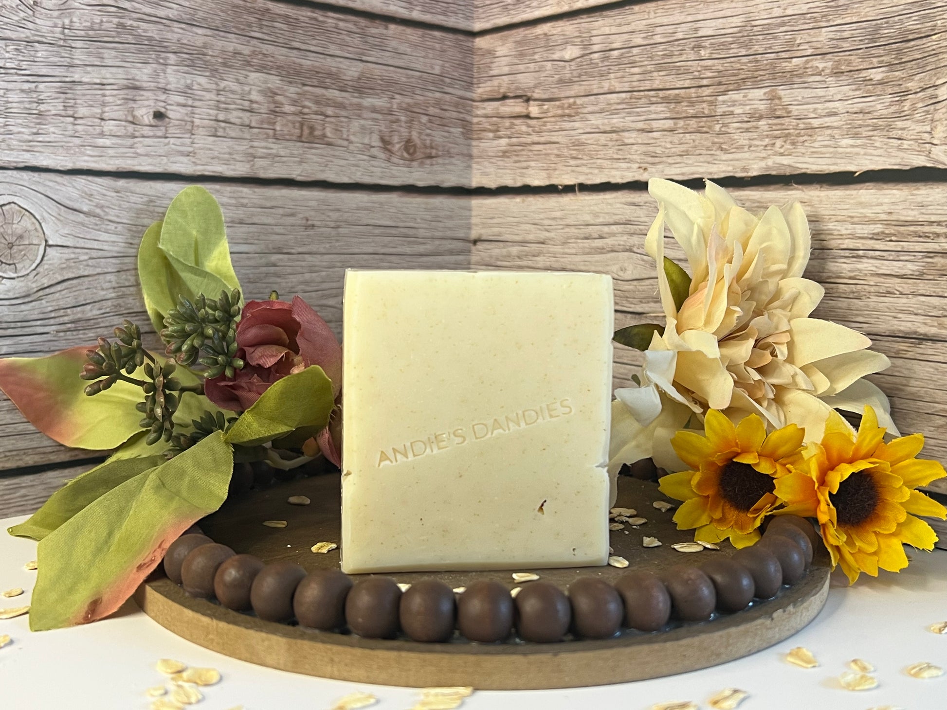 A rectangular bar of natural soap standing upright on a wooden tray, surrounded by dried flowers, sunflowers, and oats against a rustic wood plank background. The soap is embossed with the brand name 'ANDIE'S DANDIES'.