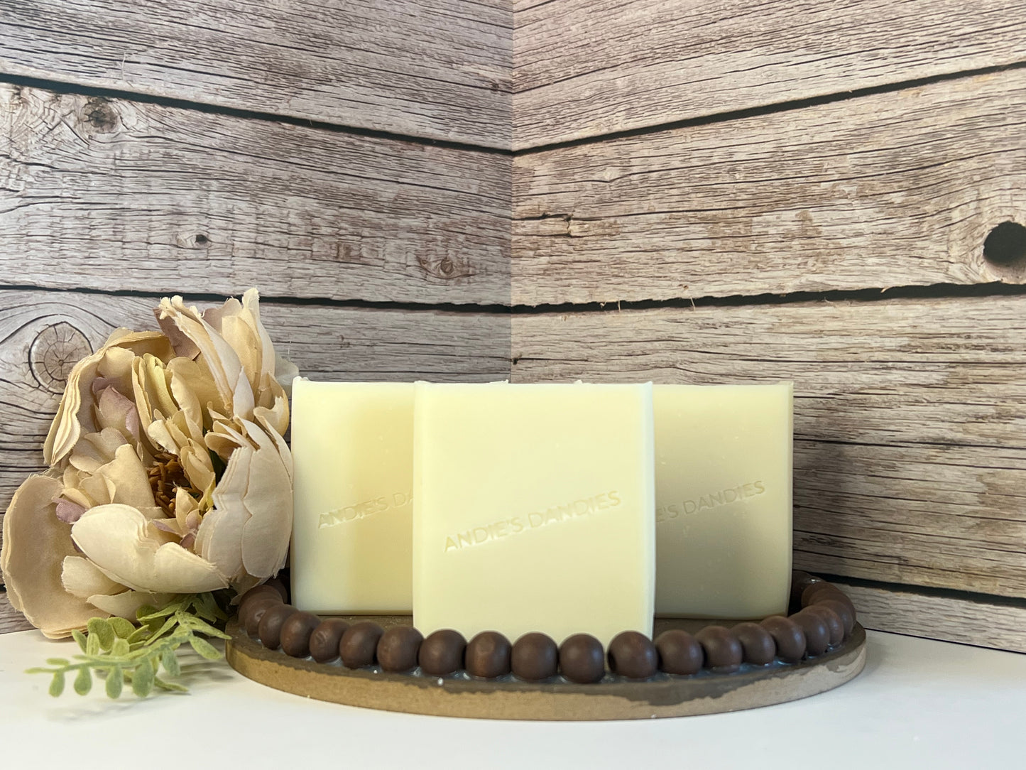 Three rectangle bars of pale cream soap standing upright on a round wooden tray with a beaded edge, styled with a large flower and green leaves on the left side, against a rustic wood plank background. The soap is embossed with the brand name 'ANDIE'S DANDIES'.