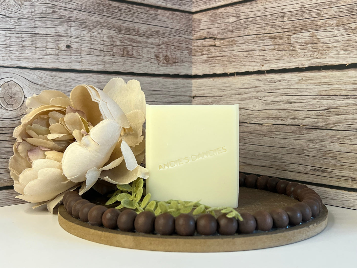 A rectangle bar of pale cream soap standing upright on a round wooden tray with a beaded edge, styled with a large flower and green leaves on the left side, against a rustic wood plank background. The soap is embossed with the brand name 'ANDIE'S DANDIES'.