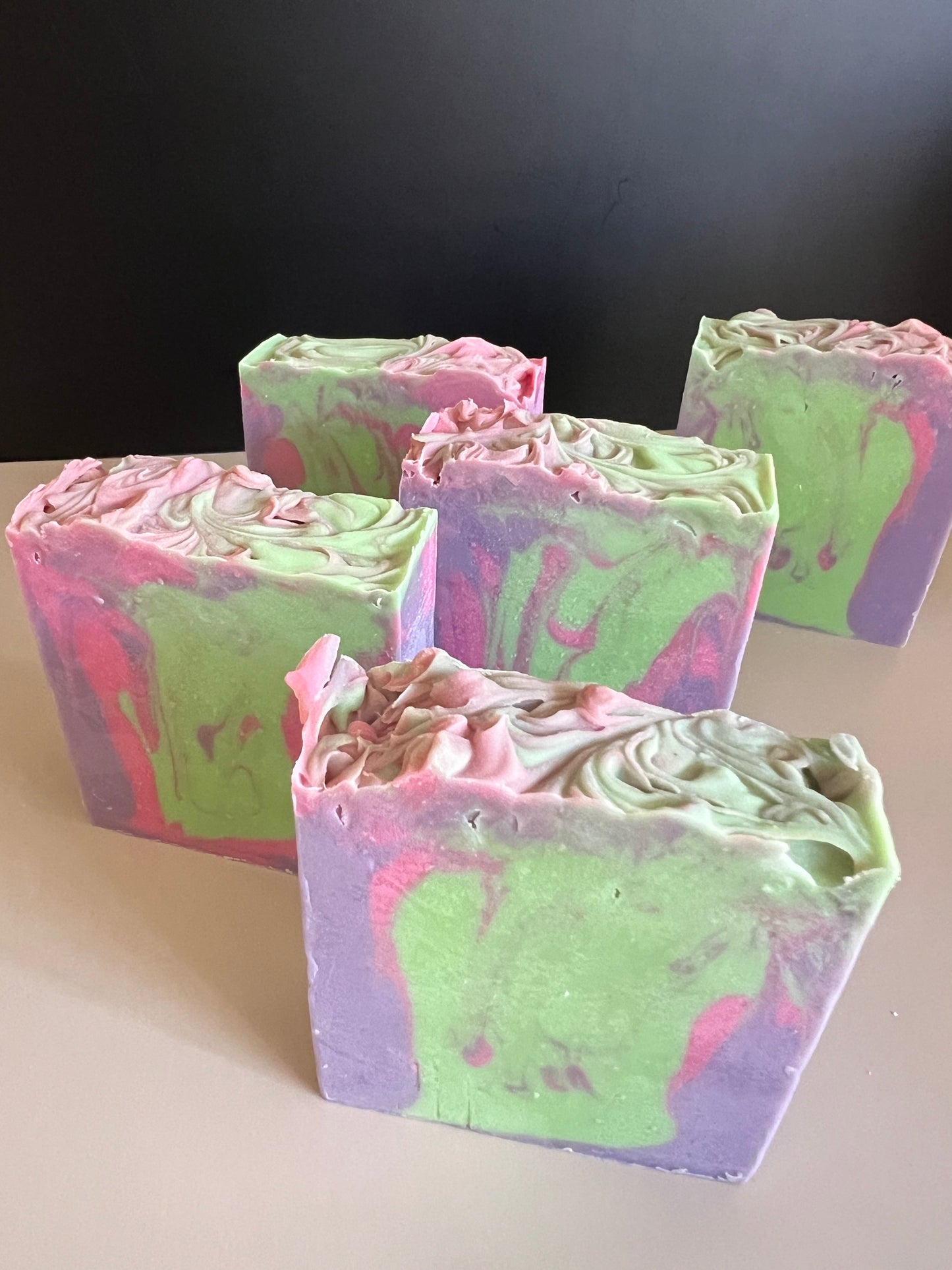 Five handmade marbled soap bars in pastel green, purple, and pink, arranged on a light beige surface against a dark gray background.