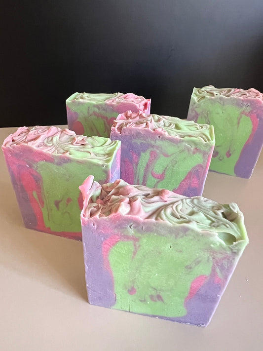 Five handmade marbled soap bars in pastel green, purple, and pink, arranged on a light beige surface against a dark gray background.