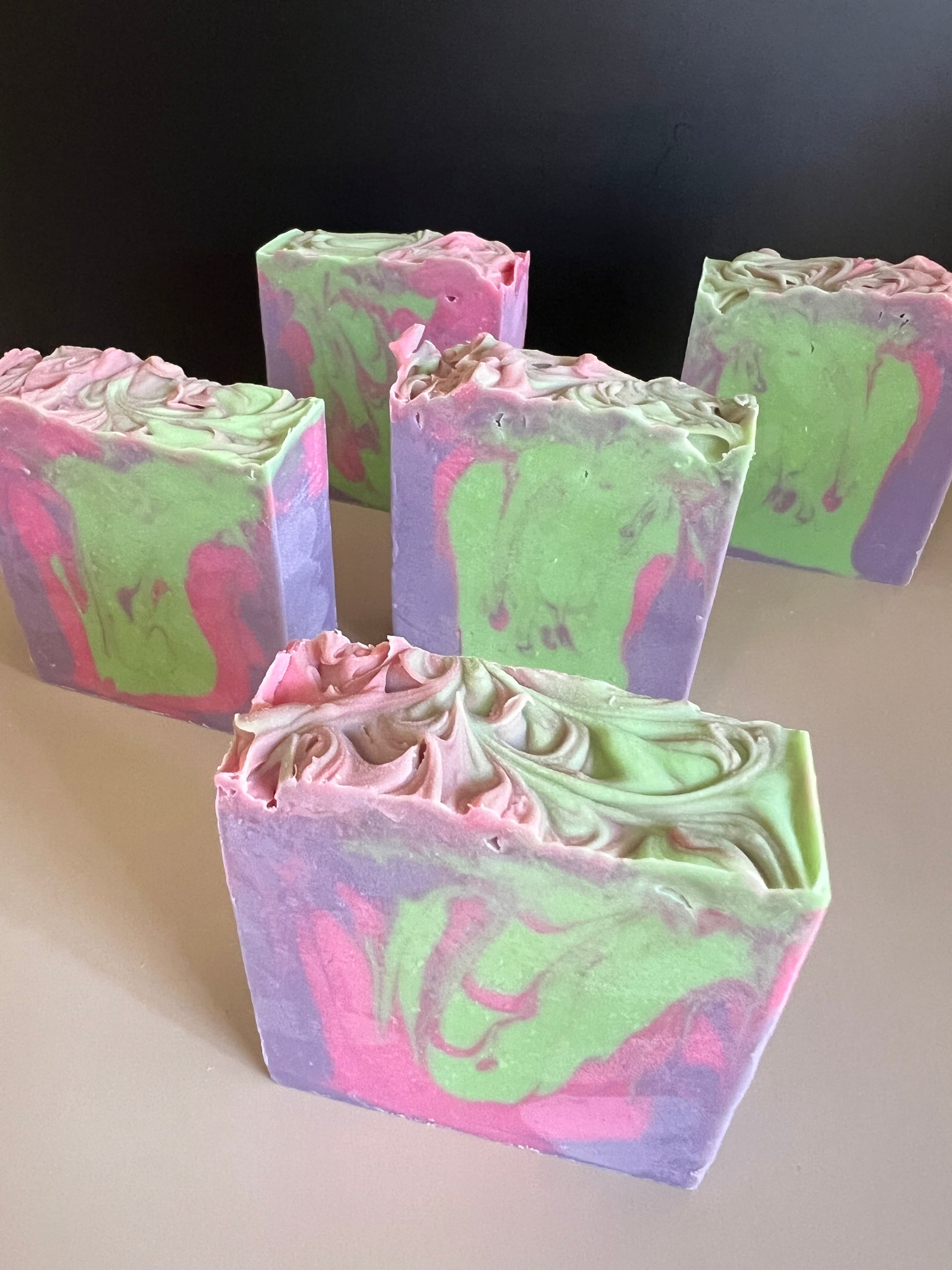 Five rectangular bars of handmade soap with swirled pink, green, and purple patterns, arranged on a light beige surface against a dark background.